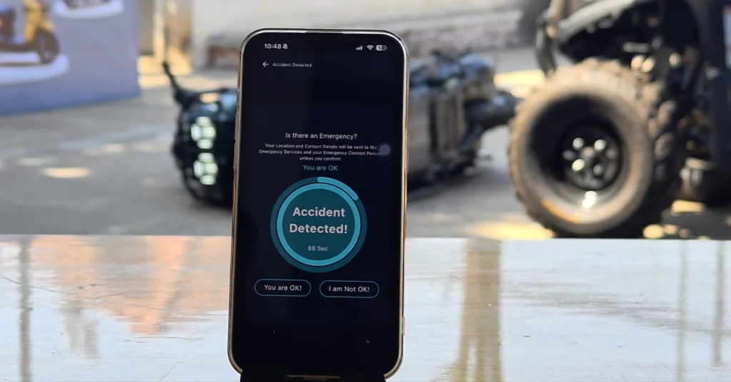 A smartphone on a table shows an accident detection alert message during a simulated crash, with a Calypso Nijia scooter on the ground in the background.